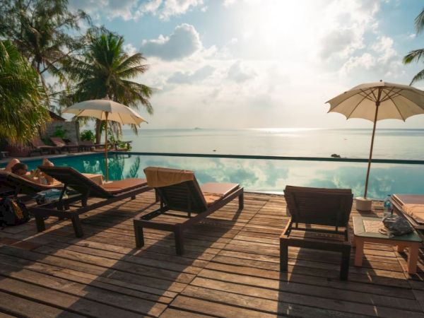 A scenic view of a poolside area with lounge chairs, umbrellas, and palm trees facing a serene body of water under a sunny sky.
