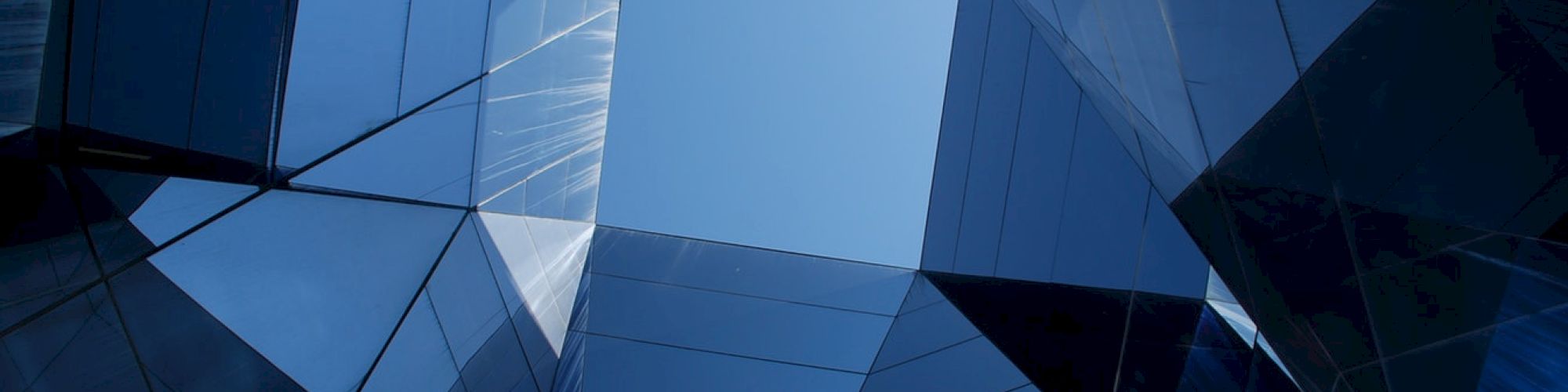 The image shows a view of a blue sky framed by modern, reflective glass architecture creating a geometric pattern.