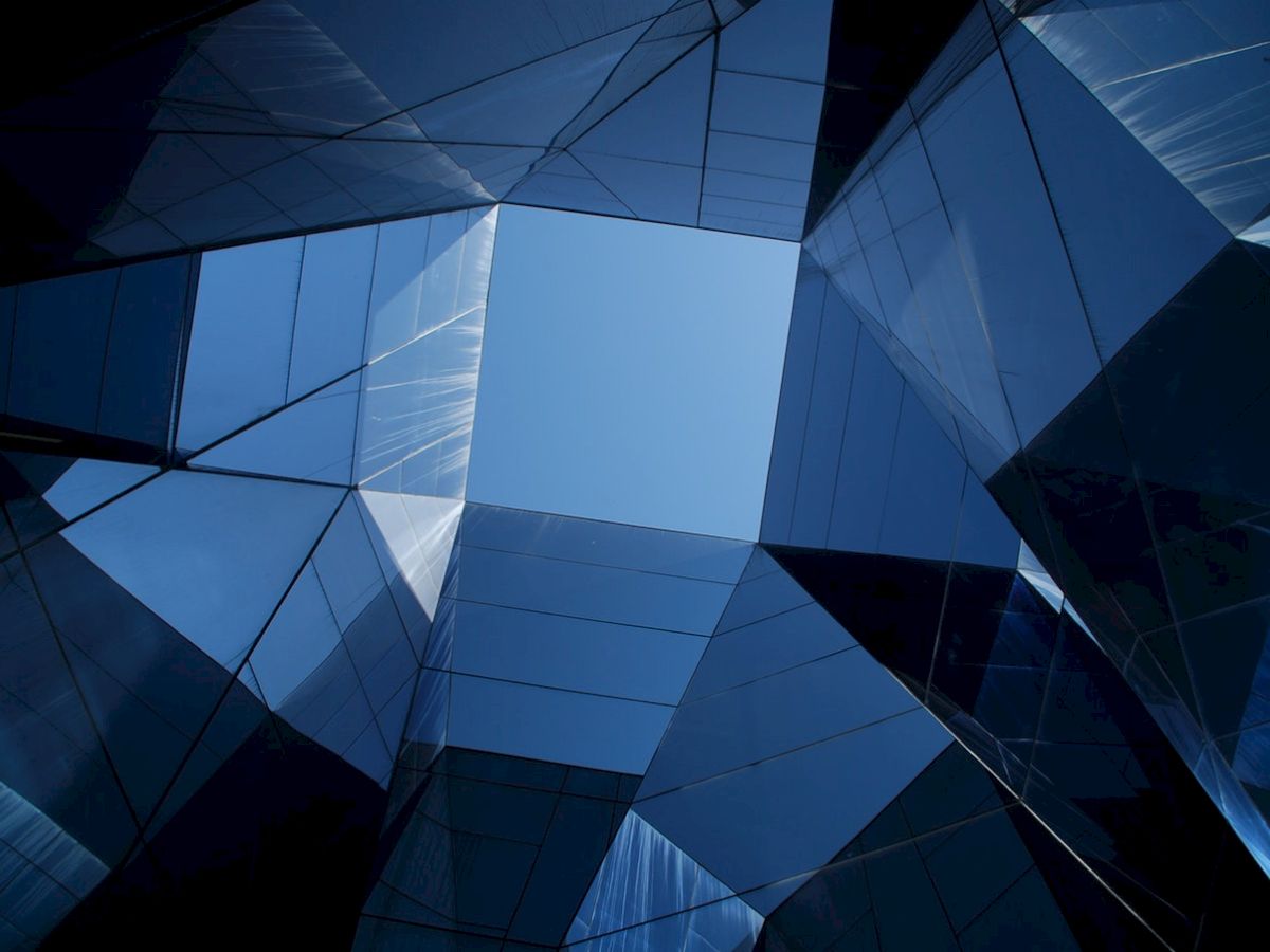 The image shows a striking architectural view looking up at a geometric arrangement of glass buildings against a clear blue sky.