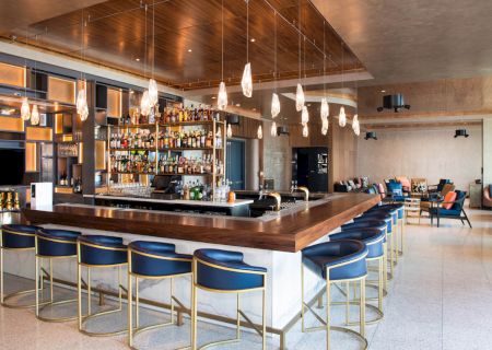 A modern bar features a central wooden counter with blue and gold stools, hanging lights, and a fully stocked display of bottles.
