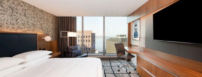 A modern hotel room with a large bed, TV, desk, and a view of city buildings through a window, featuring contemporary decor and lighting.