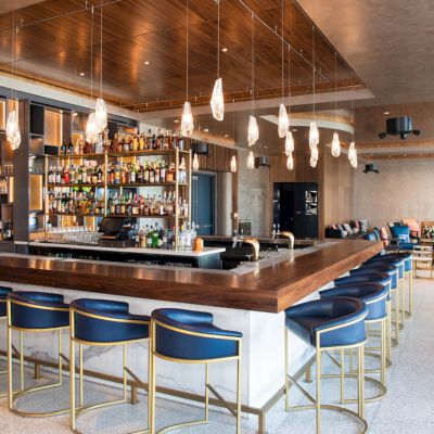 A modern bar with blue chairs, hanging lights, and a well-stocked drink display. The setting is sleek with a cozy seating area adjacent.