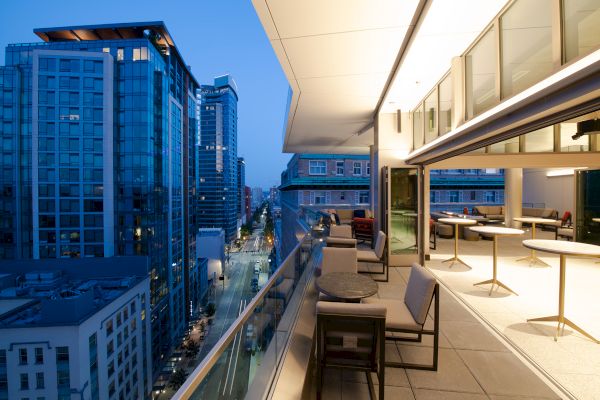 Rooftop view of modern buildings and a city street at dusk, featuring an elegant, open-air seating area with tables and chairs.