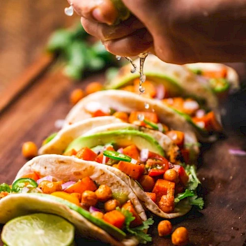 A hand squeezes lime over several tacos filled with chickpeas, vegetables, and garnished with cilantro, placed on a wooden board.