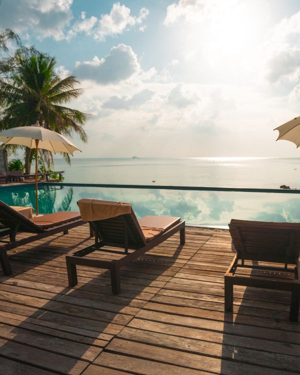 Lounge chairs and umbrellas by a pool overlook a serene ocean, surrounded by palm trees under a partly cloudy sky.