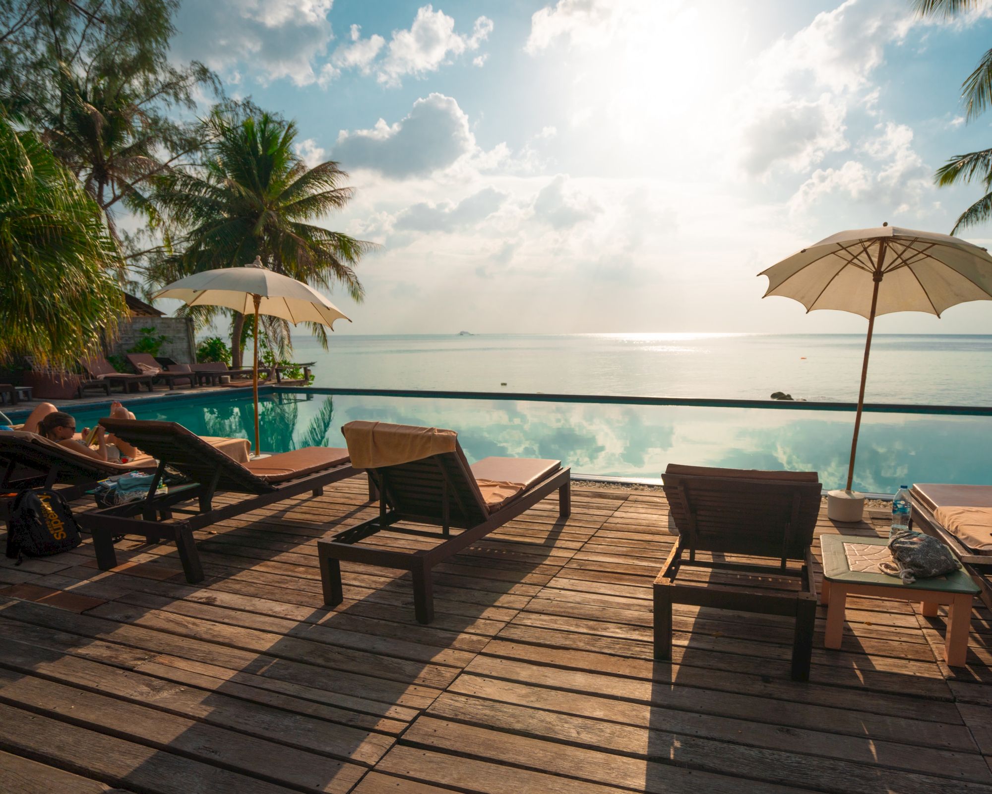 Sun loungers and umbrellas by a pool, overlooking the ocean under a bright sky with palm trees in the background.
