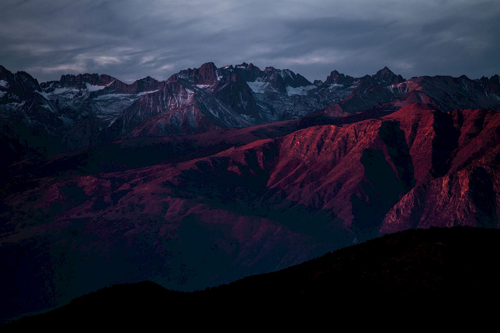 A mountain range at sunset, with peaks and ridges bathed in red hues, under a cloudy sky.