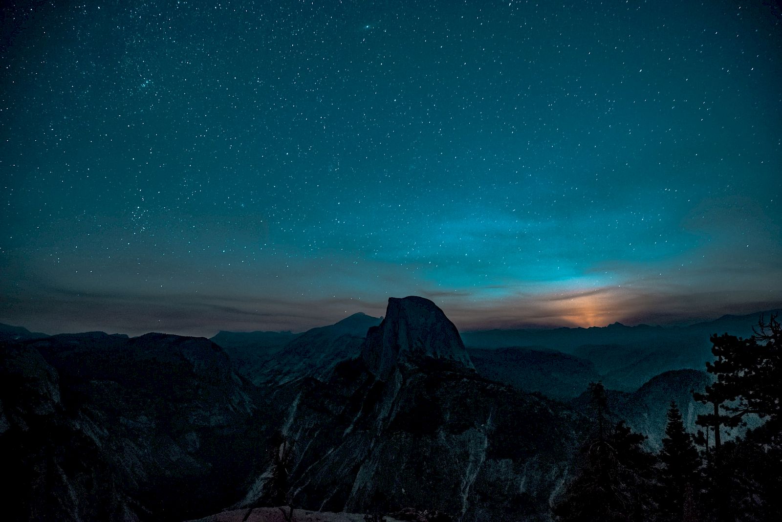A mountain landscape under a starry night sky, with a glow from a setting or rising moon behind the mountains.