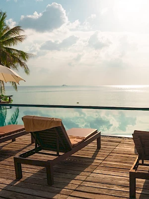 The image shows a serene poolside scene with lounge chairs, umbrellas, and a view of the ocean under a partly cloudy, sunlit sky.