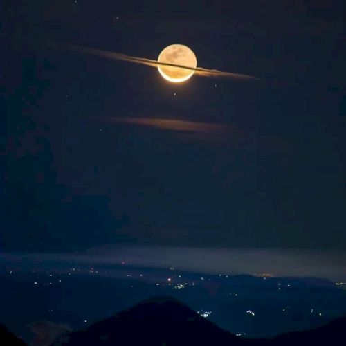 A full moon appears with horizontal clouds, resembling Saturn with its rings, above a landscape dotted with city lights.