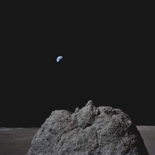 A view of Earth rising against the blackness of space, seen from the surface of the Moon with a rocky foreground.