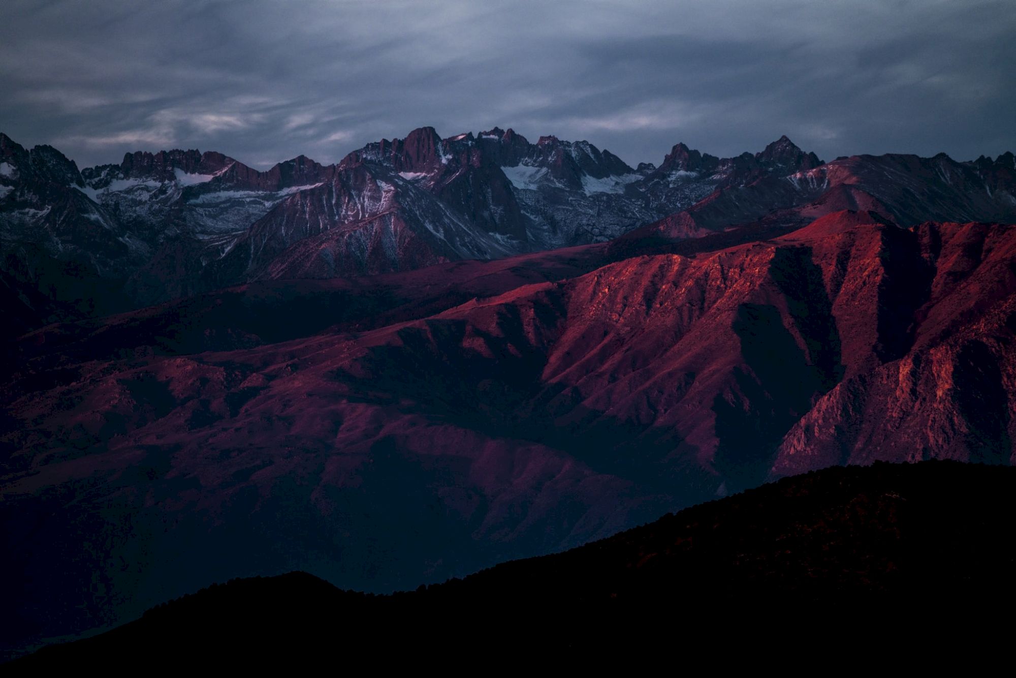 A mountain range bathed in red and purple hues during sunset, with dramatic clouds above and rugged peaks in the background.