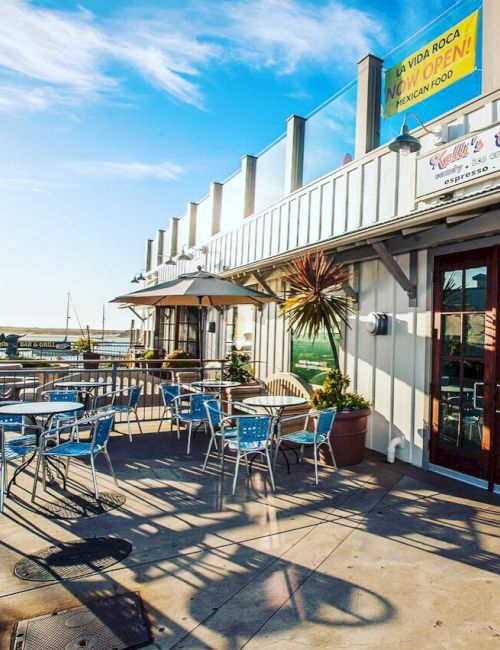 Outdoor dining area with tables and chairs by a waterfront, featuring a building with signage under a clear blue sky.