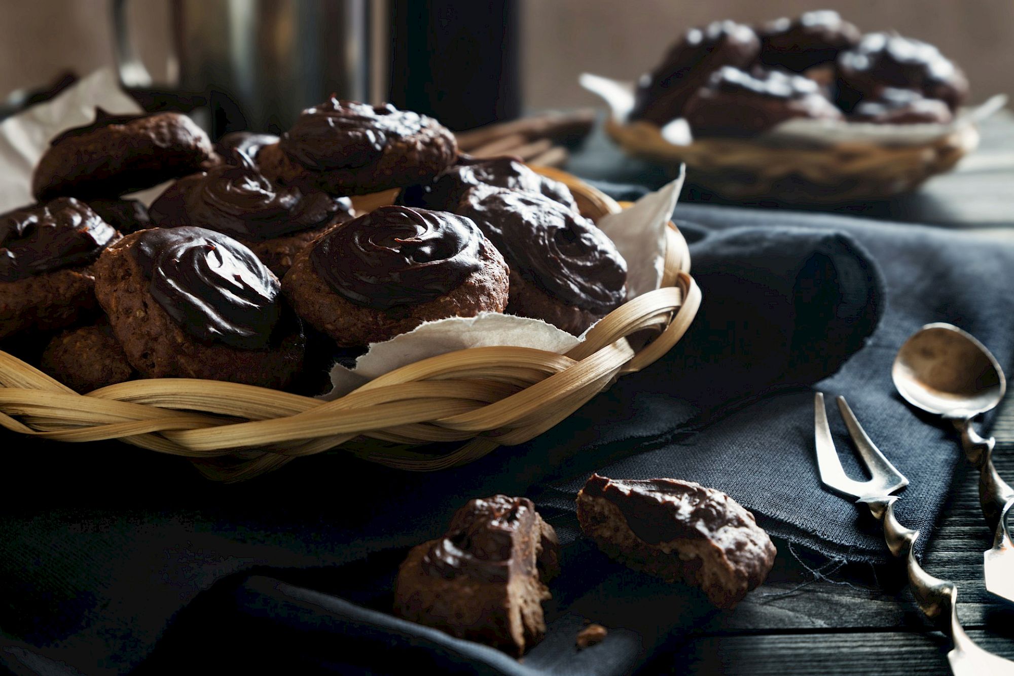 A basket of chocolate cookies with a swirl pattern, a few broken, placed on a dark cloth alongside cutlery.