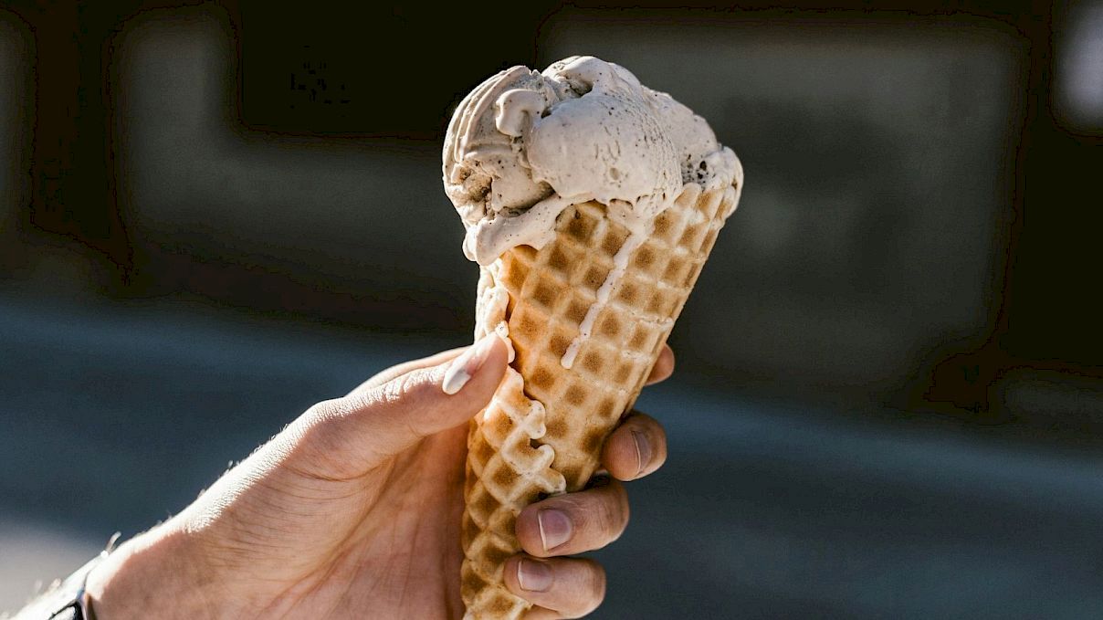 A person is holding an ice cream cone with two scoops of ice cream in a waffle cone, set against a blurred outdoor background.