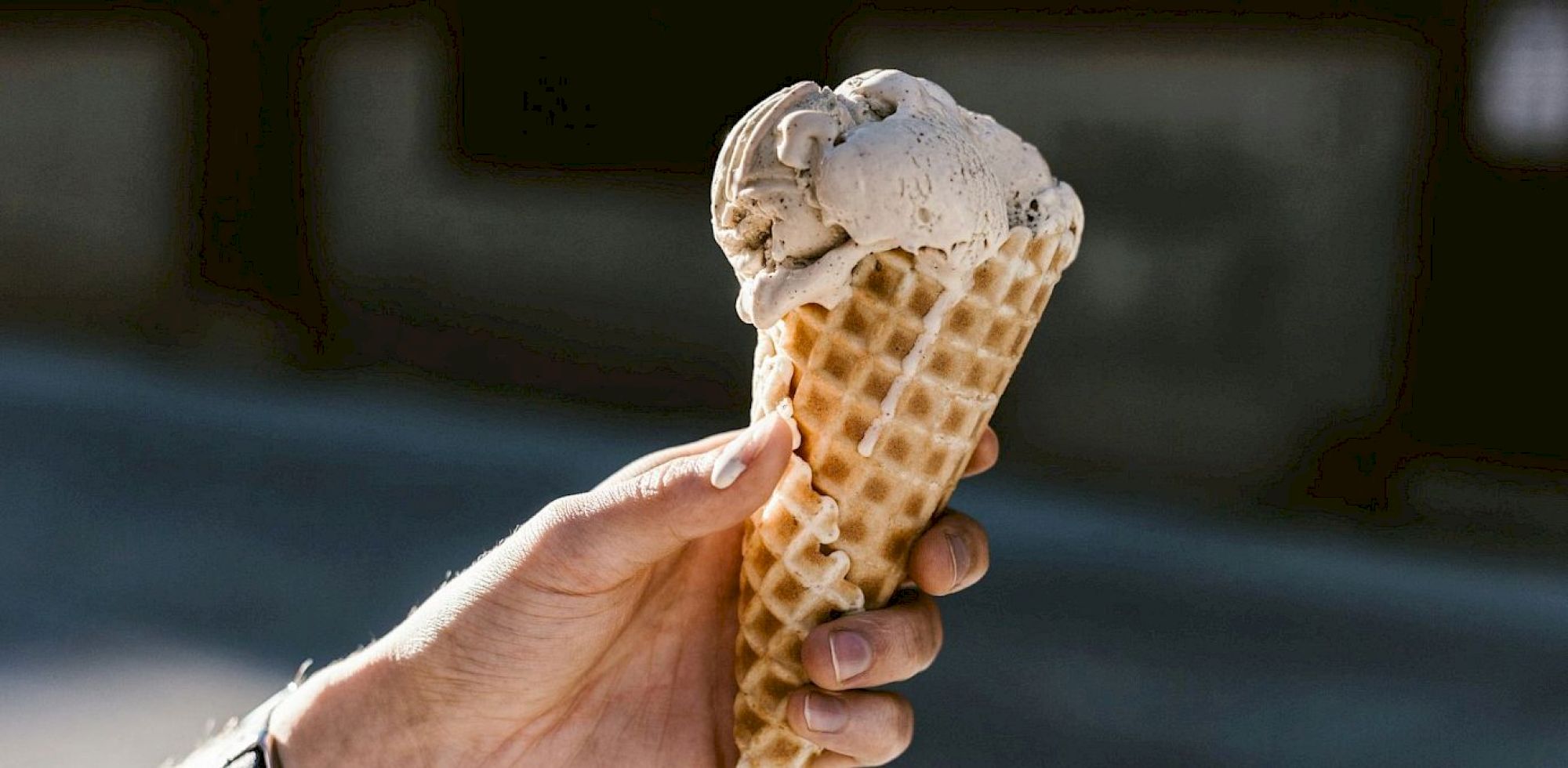 A person is holding an ice cream cone with two scoops of ice cream in a waffle cone, set against a blurred outdoor background.