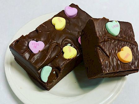 Two chocolate fudge pieces topped with colorful heart-shaped candies on a white plate, set against a plain background.