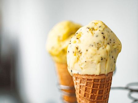 Two waffle cones with yellow ice cream and small specks, likely fruit or seeds, in a soft-focus background.