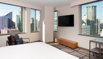 A modern hotel room with city views, featuring a bed, wall-mounted TV, armchair, and windows showing a skyline.