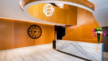 A modern lobby with wooden accents, a large clock, a marble reception desk, and decorative lighting overhead.