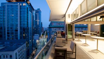 A modern balcony with seating overlooks a cityscape of tall buildings and lit streets during the evening.