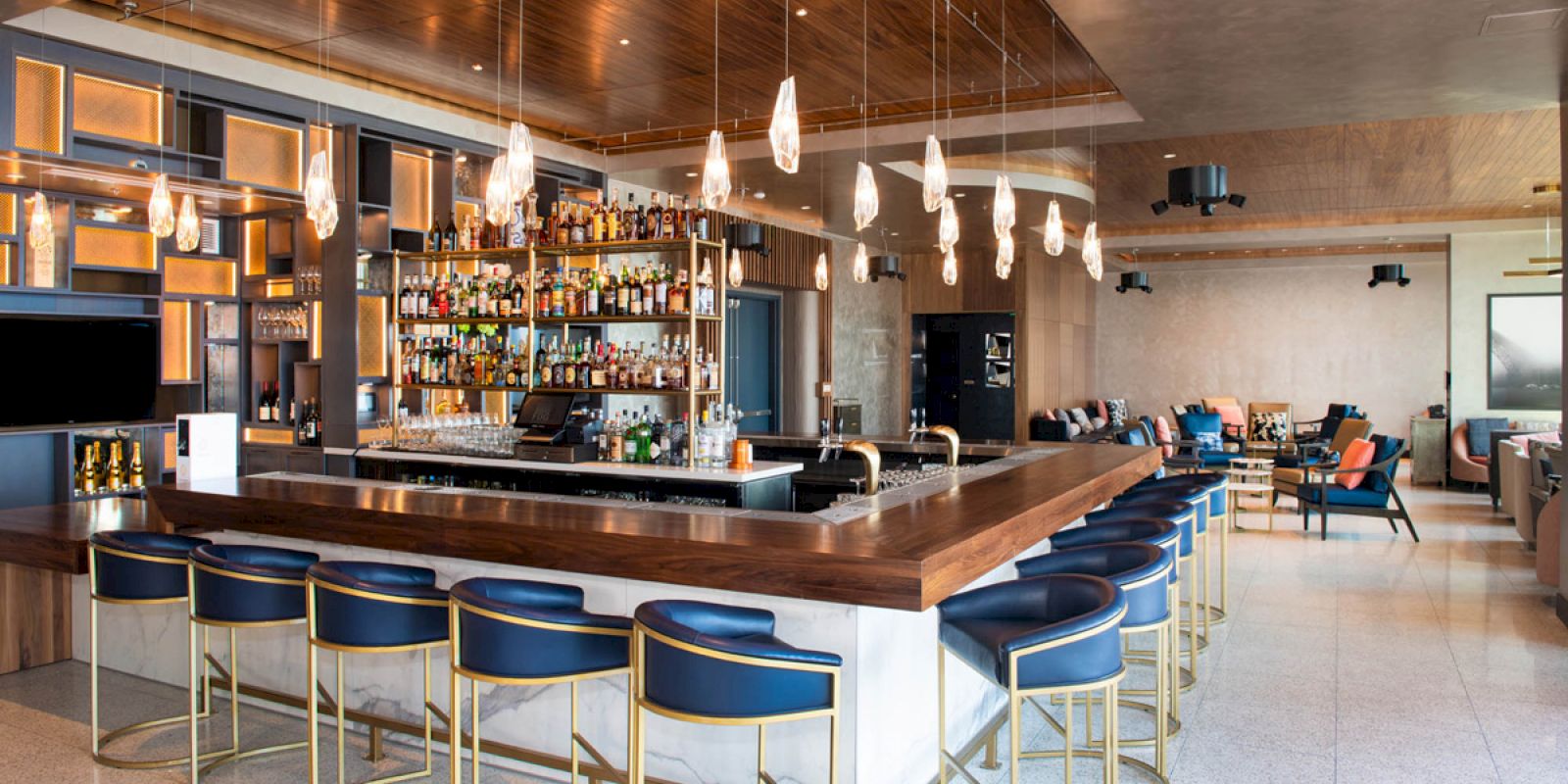 A modern bar with a wooden counter, blue stools, and hanging lights. Shelves stocked with bottles are behind the counter, creating a cozy atmosphere.