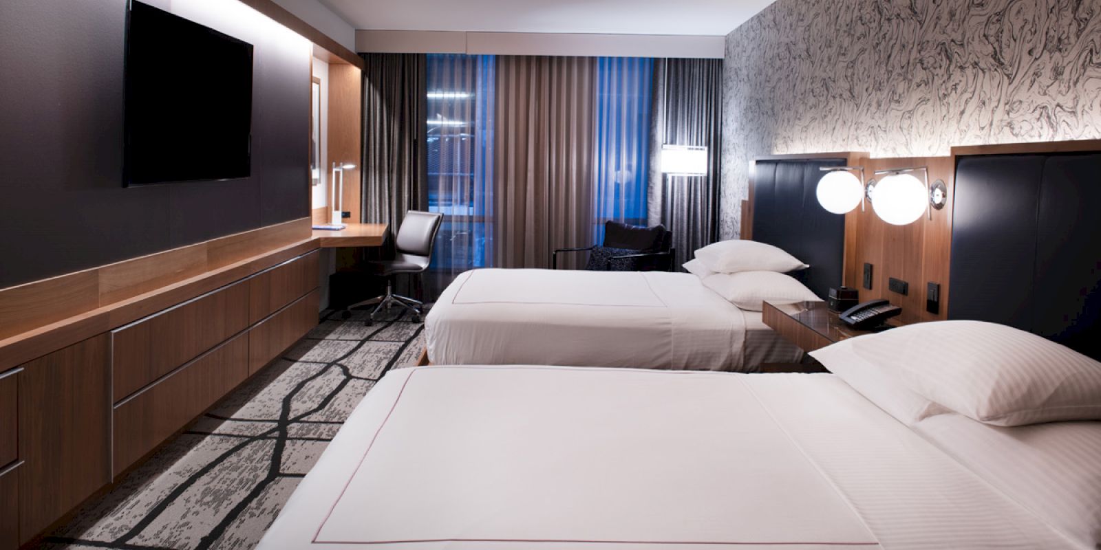 The image shows a modern hotel room with two neatly made beds, a wall-mounted TV, a desk with a chair, and stylish lighting.