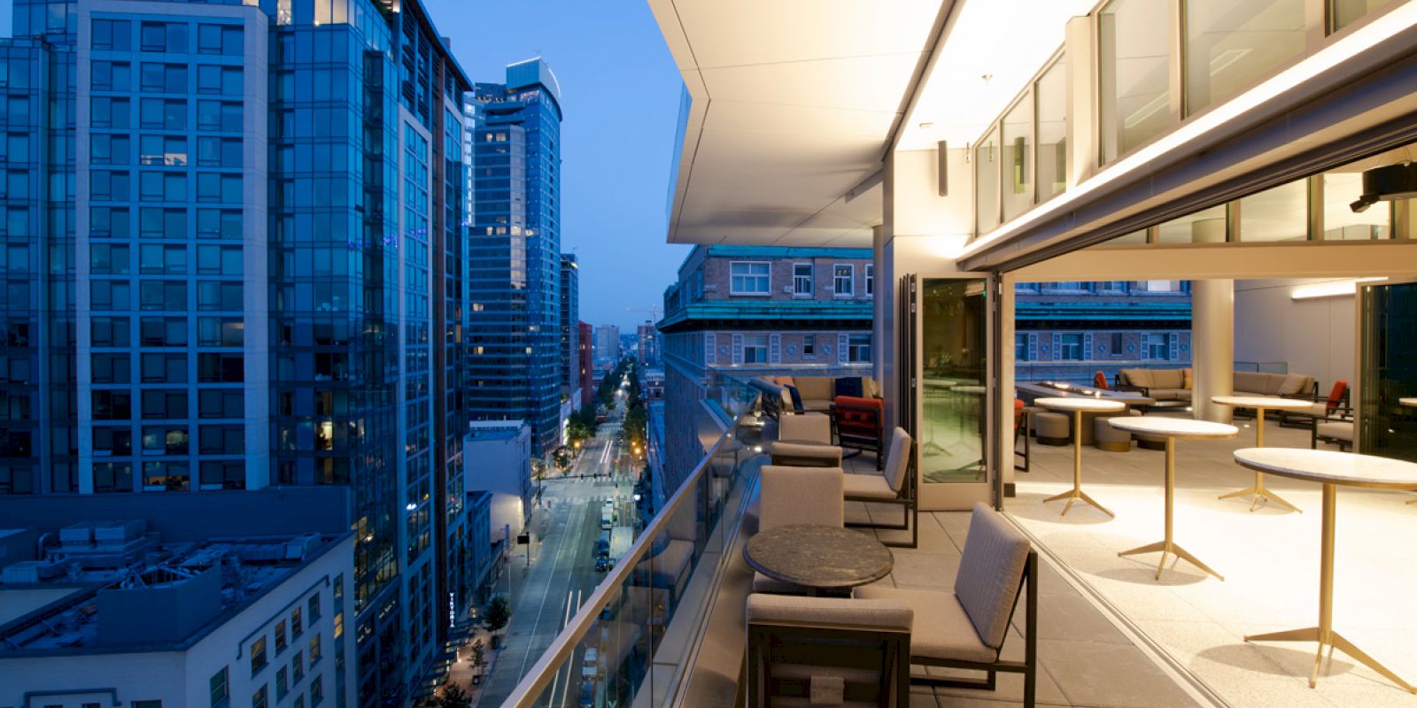 A modern cityscape at dusk with a high-rise balcony view, featuring outdoor seating and illuminated buildings lining the street.