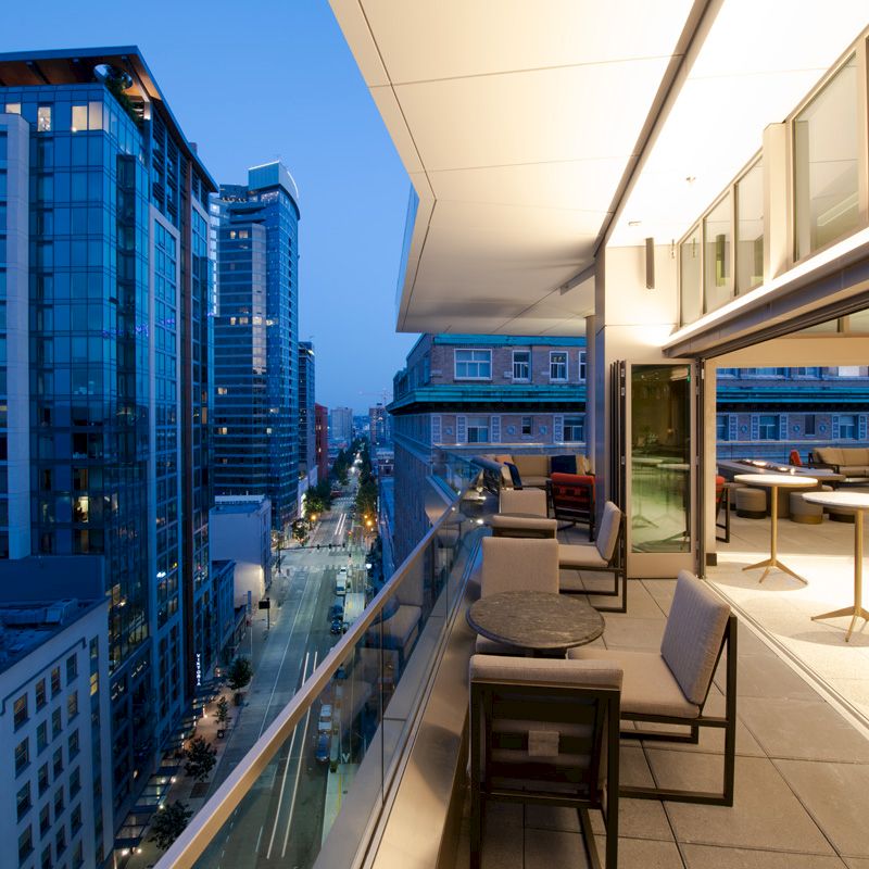 A modern rooftop terrace with seating overlooks a cityscape of tall, illuminated buildings during dusk.