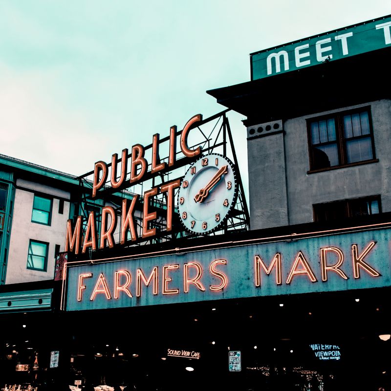 A famous public market sign with neon lights and a clock, indicating a bustling farmers market in a city setting.