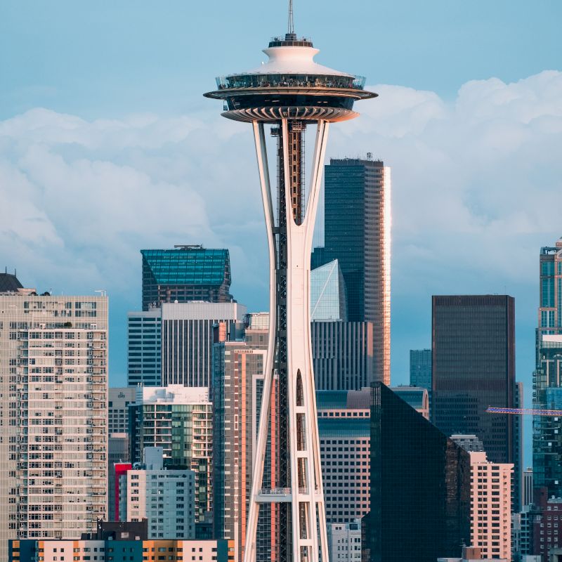 The image shows the Space Needle with surrounding skyscrapers in a city skyline under a partly cloudy sky.