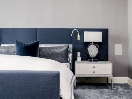 A modern bedroom with a navy blue bed, white and gray bedding, wall-mounted reading light, and a bedside table with a lamp and decorative item.