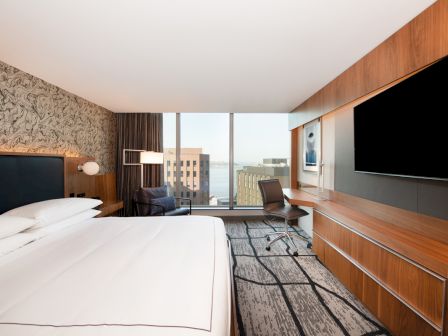 A modern hotel room with a large bed, TV, desk, and a window view of buildings and the water.