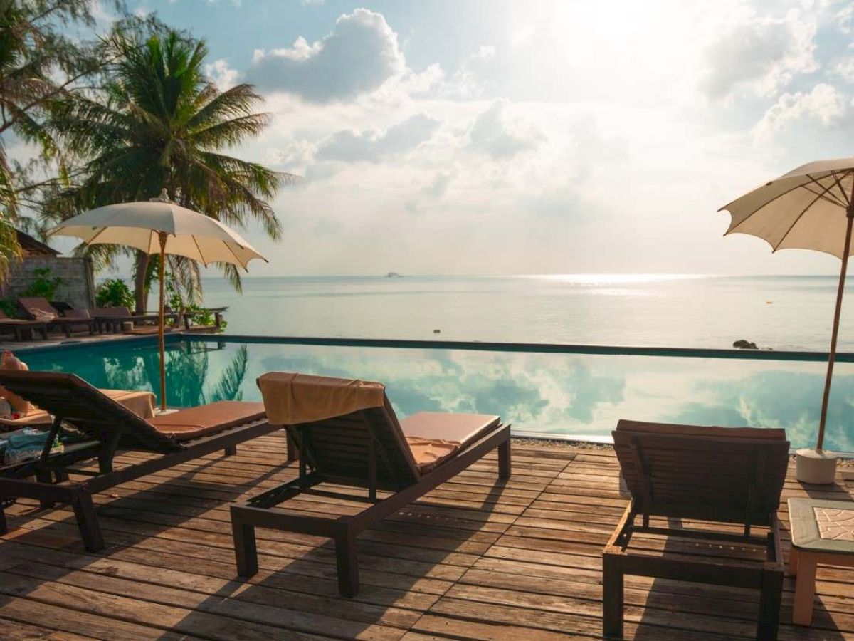 A beachfront scene with lounge chairs and umbrellas by a pool, overlooking the ocean with palm trees and a sunny sky in the background.