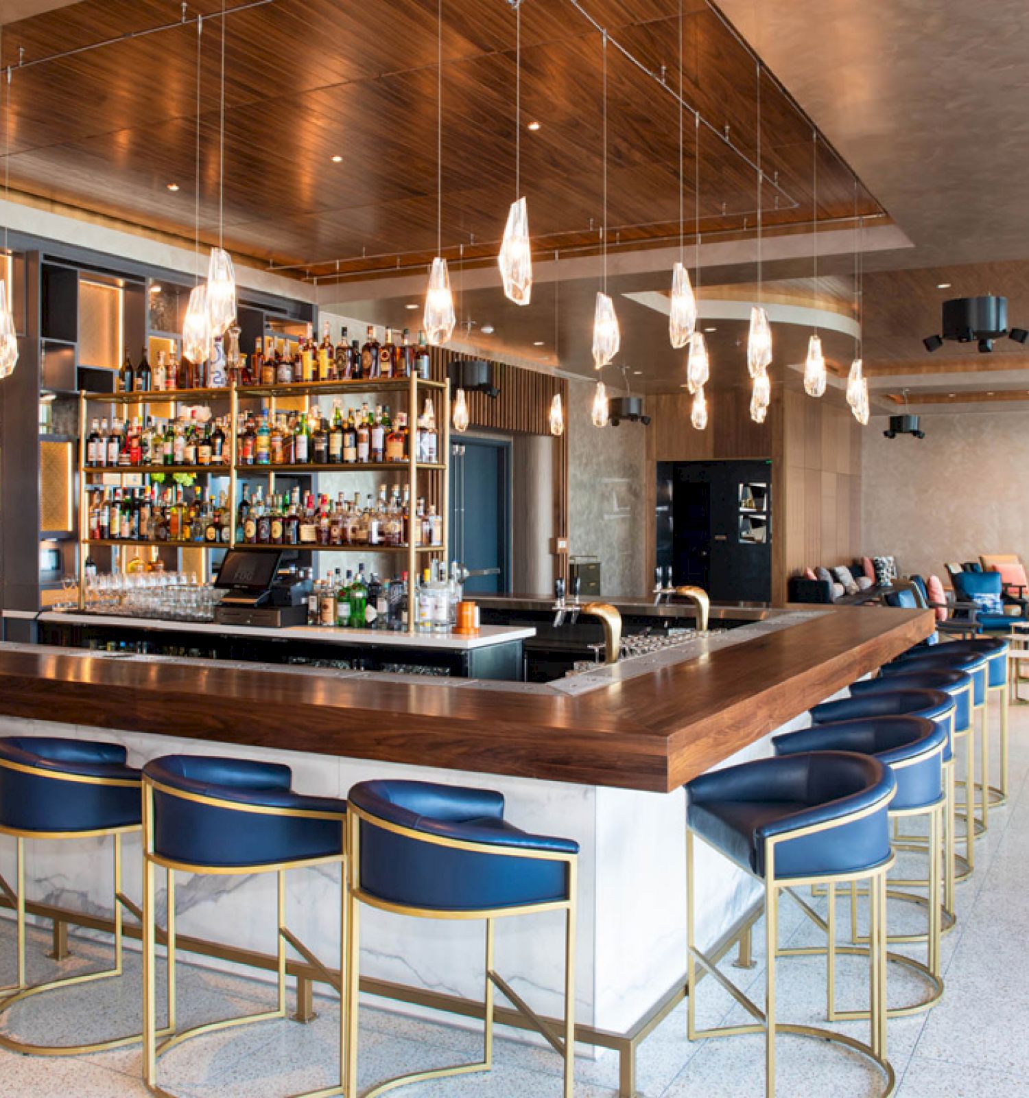 The image shows a modern bar with blue stools, hanging lights, and a well-stocked liquor shelf in a stylish interior setting.