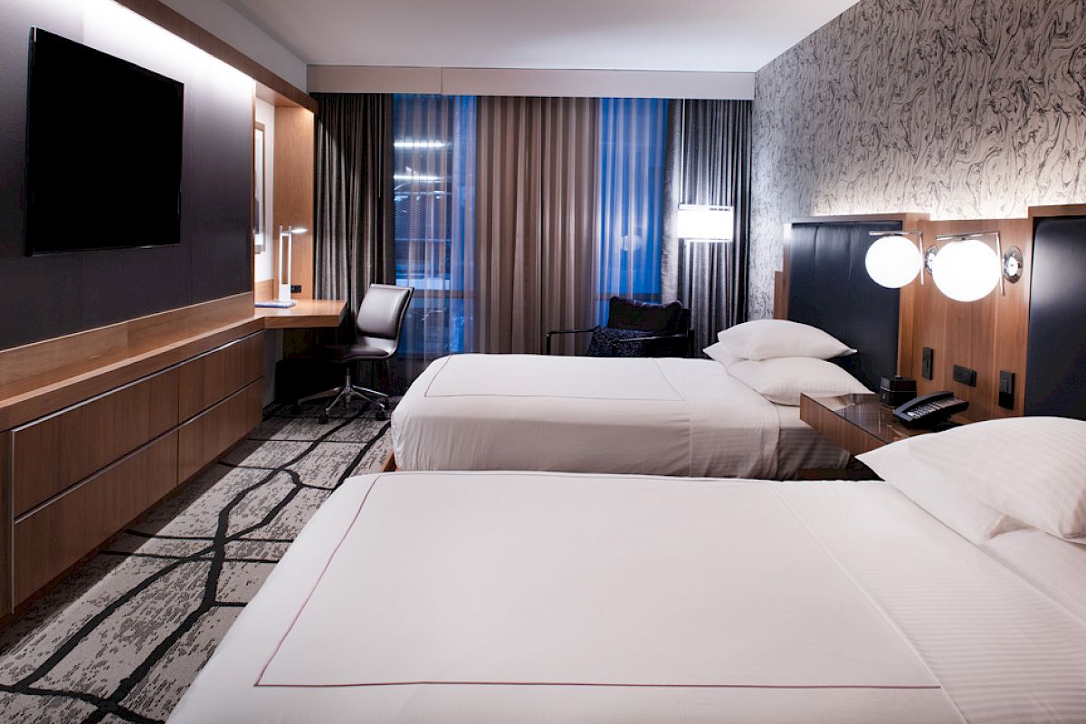 The image shows a modern hotel room with two beds, a desk, a chair, a wall-mounted TV, and stylish lighting and decor.