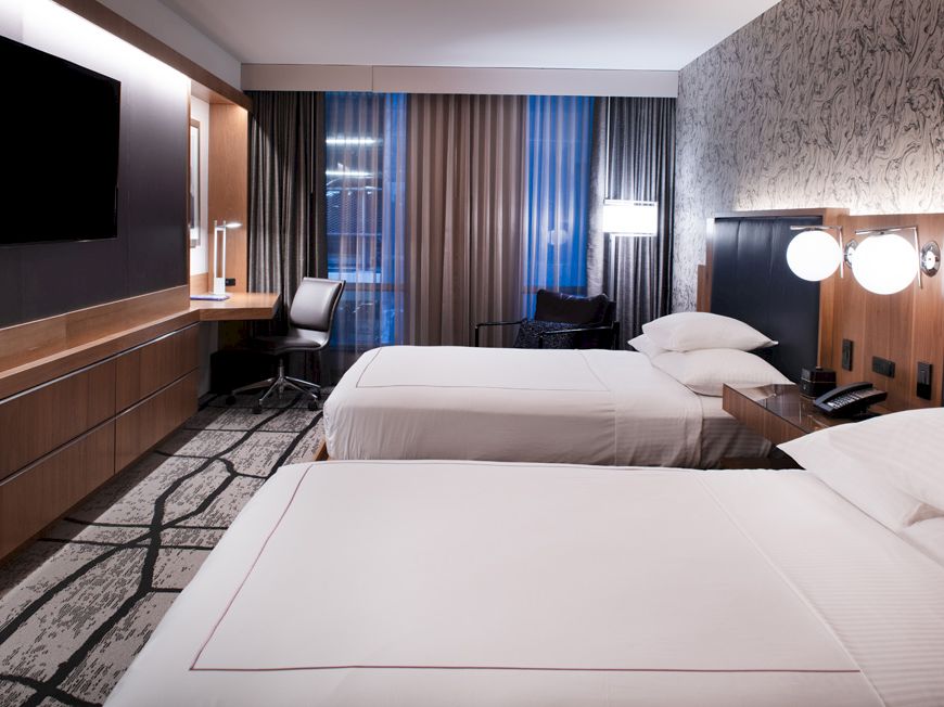 The image shows a modern hotel room with two beds, a desk, a chair, a wall-mounted TV, and stylish lighting and decor.