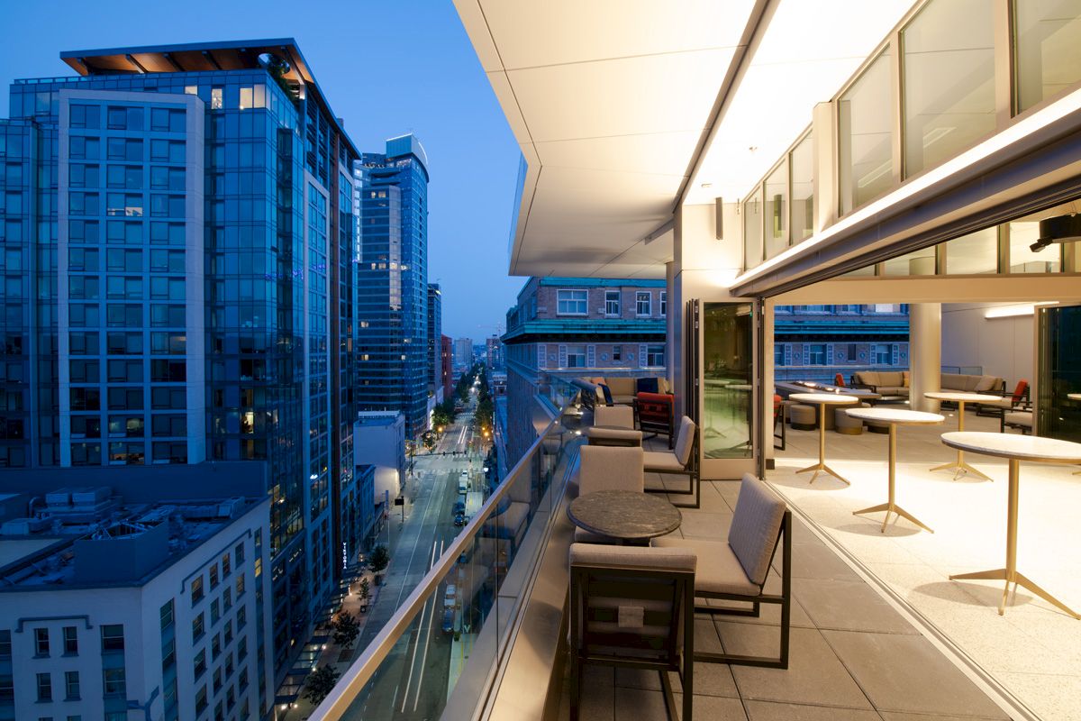 A modern balcony view overlooking a city street lined with tall buildings during dusk, with tables and chairs on the terrace.