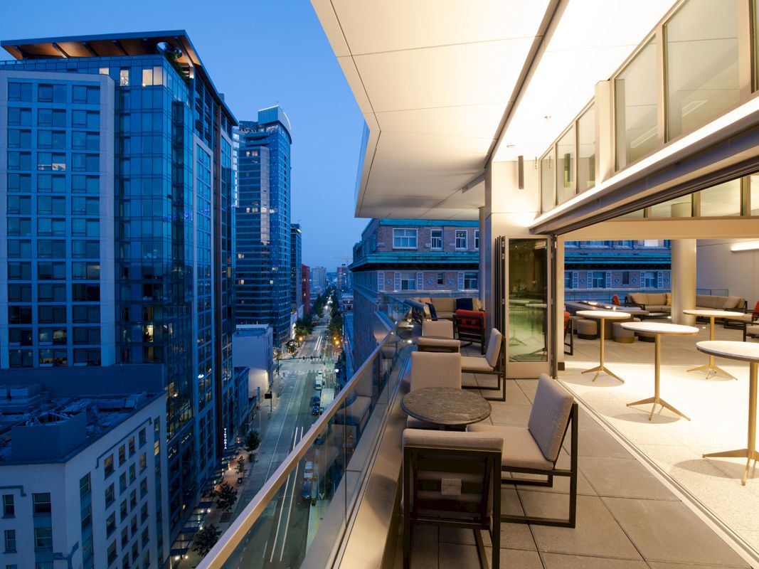 A modern balcony view overlooking a city street lined with tall buildings during dusk, with tables and chairs on the terrace.