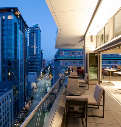 A modern rooftop terrace with seating overlooks a city street lined with tall buildings during twilight, creating an urban ambiance.