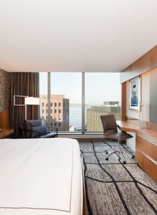 A modern hotel room with a large bed, desk, chair, and wall-mounted TV. The window offers a view of buildings and water outside.