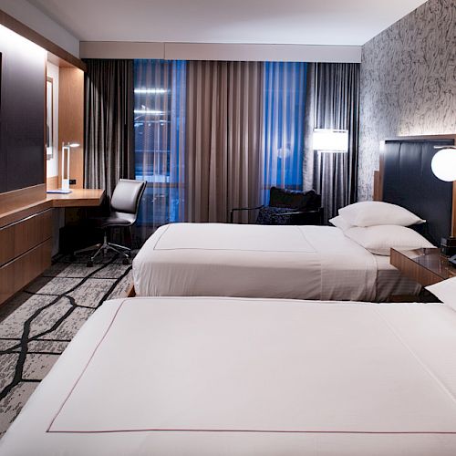 The image shows a modern hotel room with two neatly made beds, a wall-mounted TV, a desk with a chair, and stylish lighting.