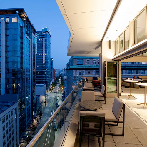 A modern balcony with glass railings overlooks a cityscape at dusk, featuring tables, chairs, and illuminated interior spaces.