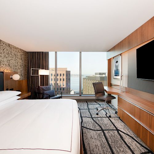 A modern hotel room features a large bed, wall-mounted TV, desk, chair, and scenic city view through expansive windows.