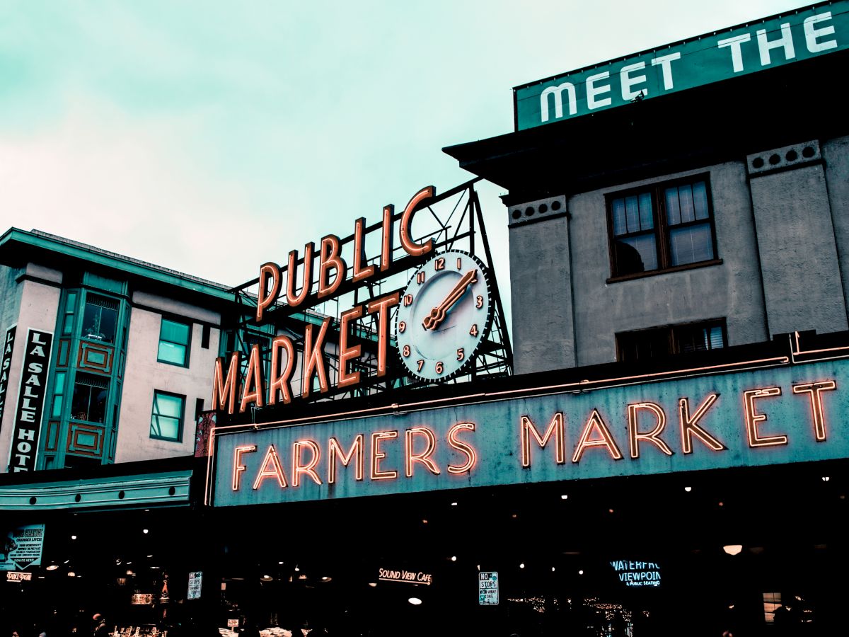The image shows a public market sign with a clock, neon "Farmers Market" text, and surrounding buildings in an urban setting.