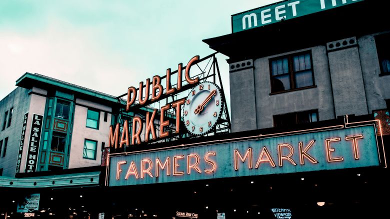 The image shows a public market sign with a clock, neon "Farmers Market" text, and surrounding buildings in an urban setting.