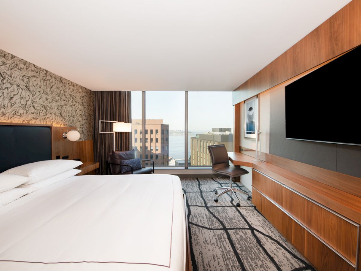 A modern hotel room with a large bed, a work desk, a wall-mounted TV, and a window view of a cityscape outside.