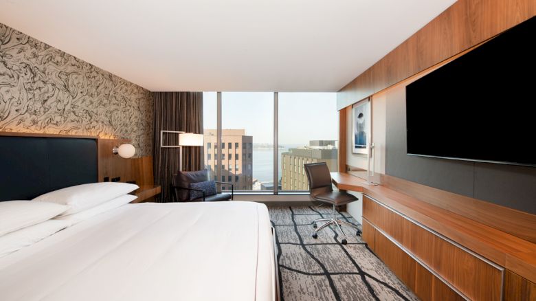 A modern hotel room with a large bed, a work desk, a wall-mounted TV, and a window view of a cityscape outside.