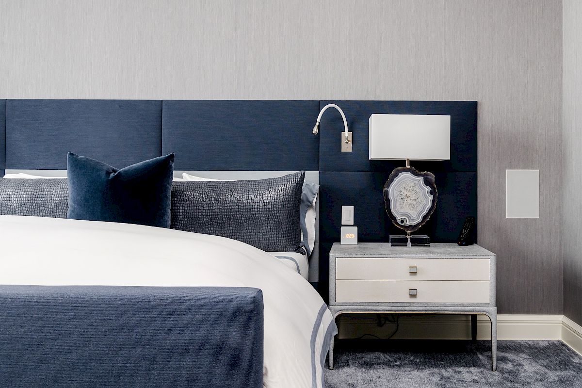The image shows a modern bedroom with a navy blue headboard, white and navy bedding, a nightstand with a lamp, and a plush carpet.