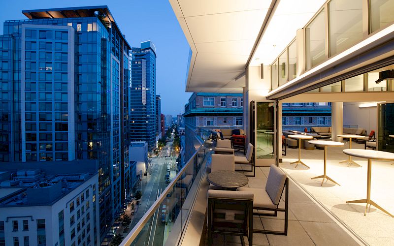 A modern city view at twilight with a sleek balcony featuring tables and chairs, overlooking illuminated high-rise buildings and a street.
